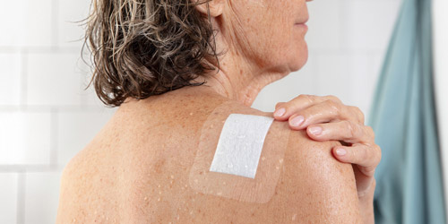 Nous voyons le dos d’une femme plus âgée. Elle porte un large bandage sur son omoplate. Nous voyons le dos d’une femme plus âgée. Elle porte un large bandage sur son omoplate.